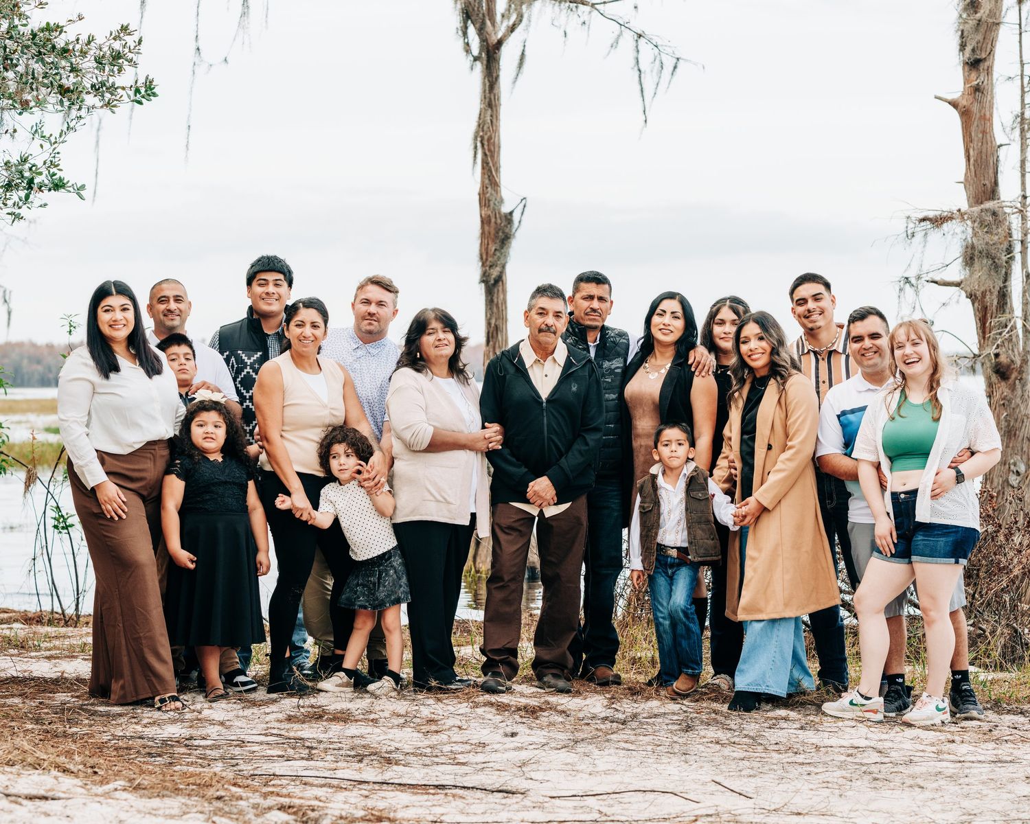 Large Hispanic family posing together outdoors near a lake, smiling for a group portrait photo.