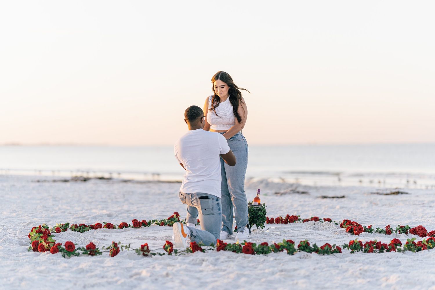Man proposing on one knee inside a rose petal heart on a white sand beach at sunset.