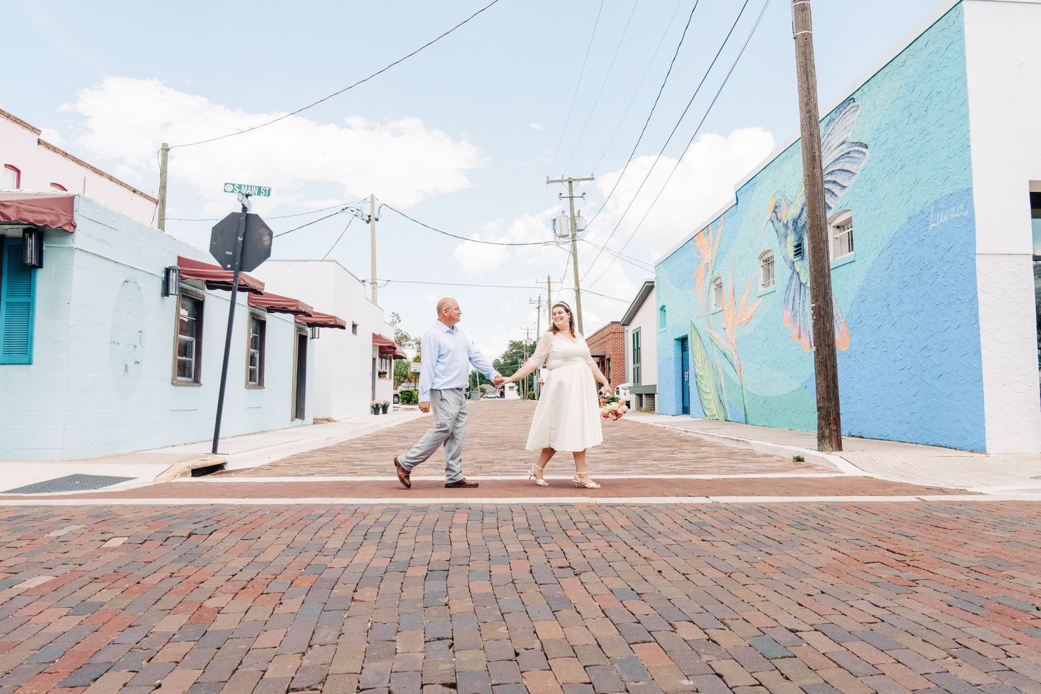 Couple holding hands walking across a brick street intersection with colorful murals and buildings in a charming urban setting.