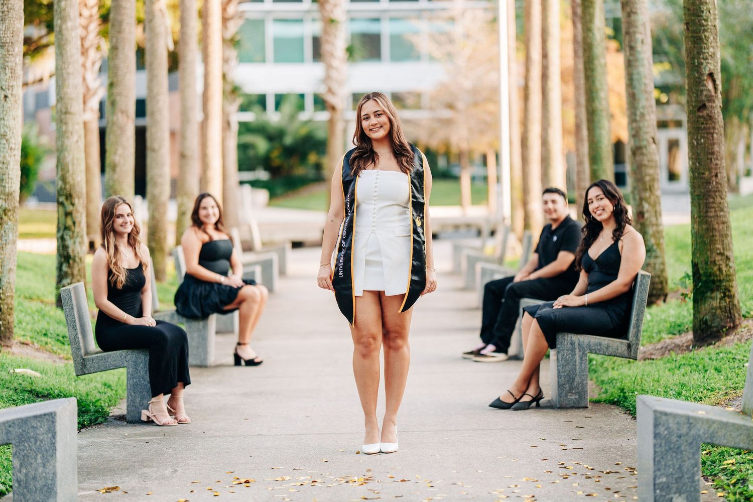 A smiling young woman in a white dress stands on a palm-lined path while friends in black outfits sit on benches behind her.