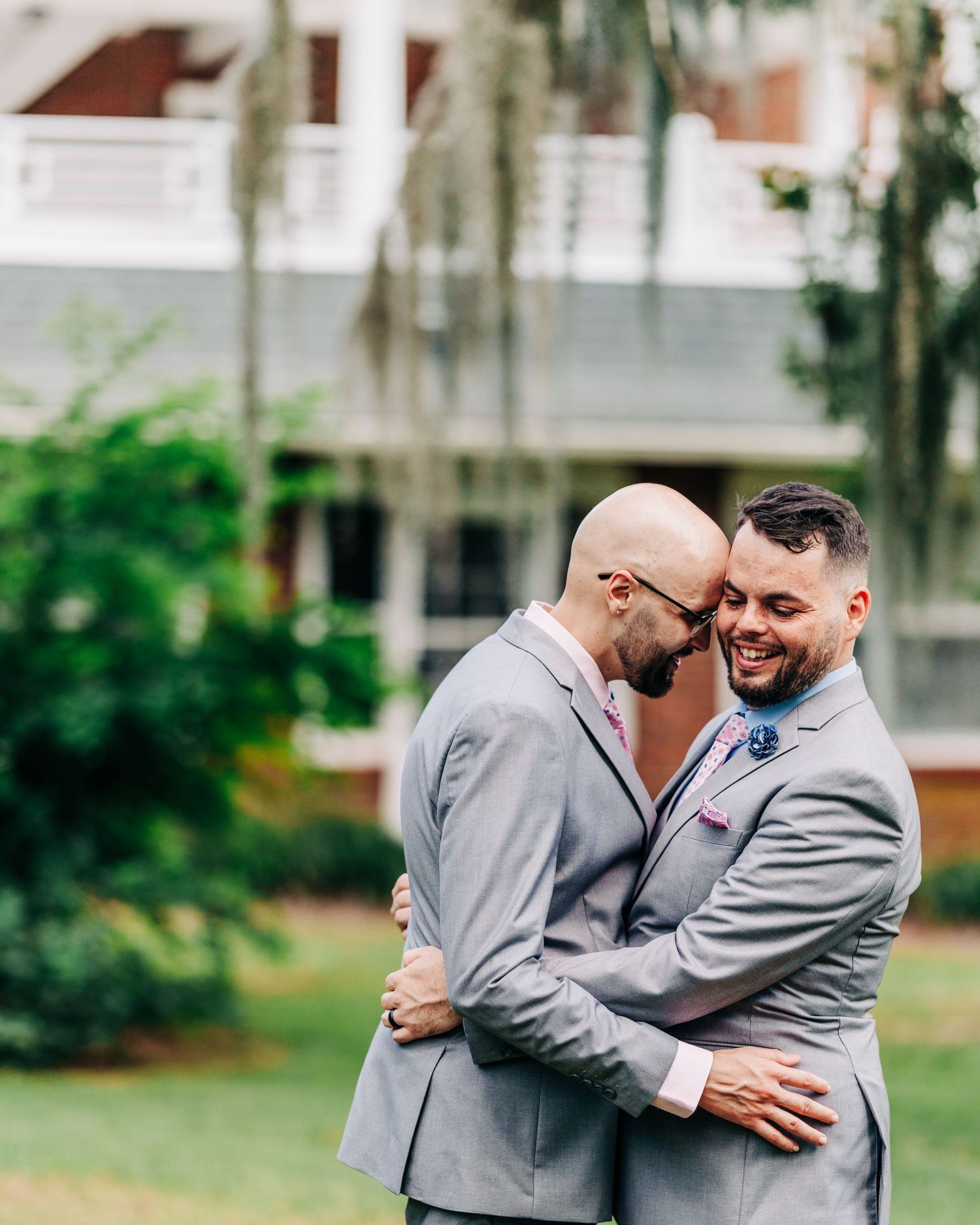 Two people in matching light gray suits share an intimate embrace while smiling outdoors in a garden setting.