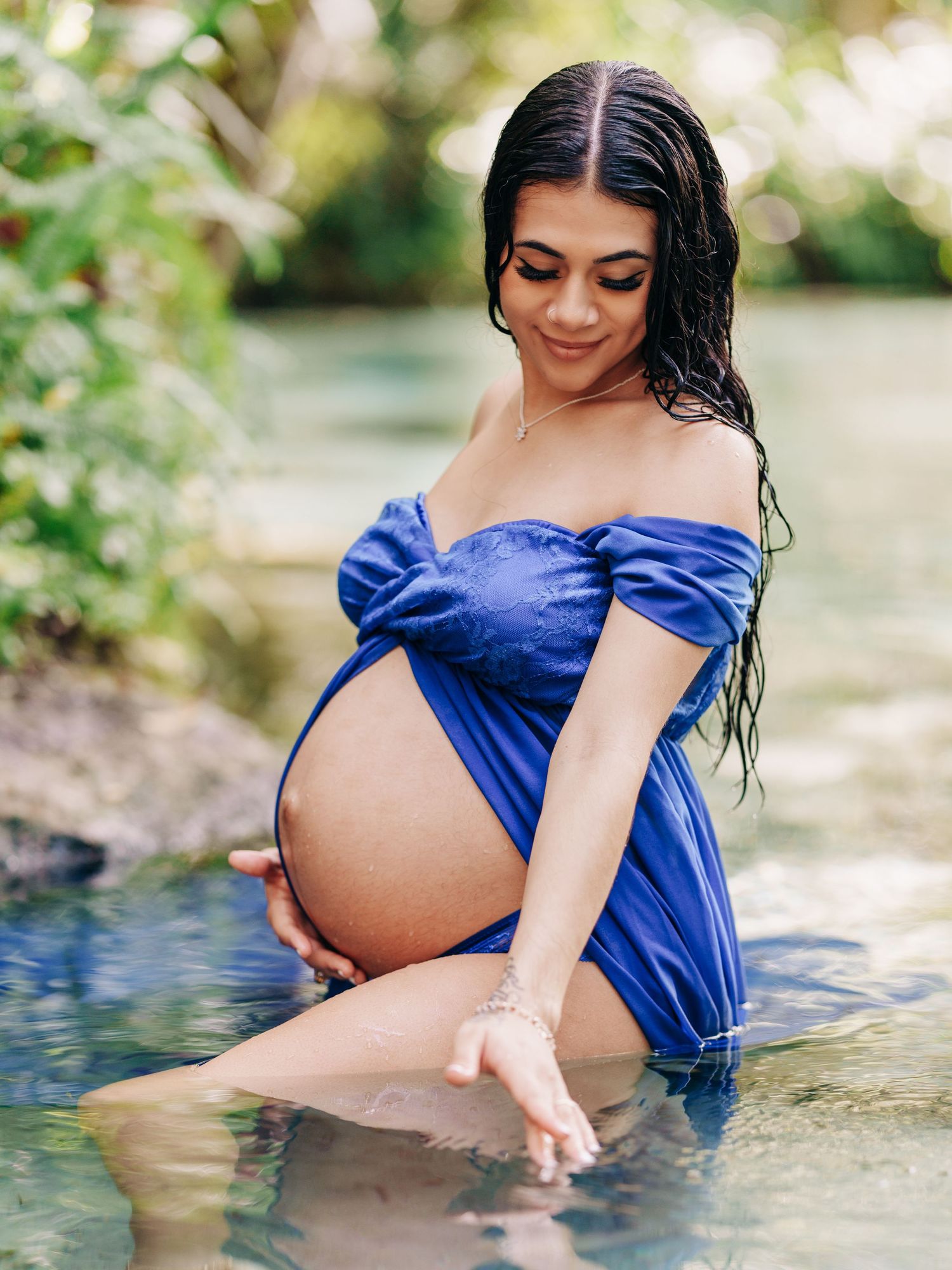 A pregnant woman in a royal blue off-shoulder dress sits in shallow water during a maternity photoshoot.