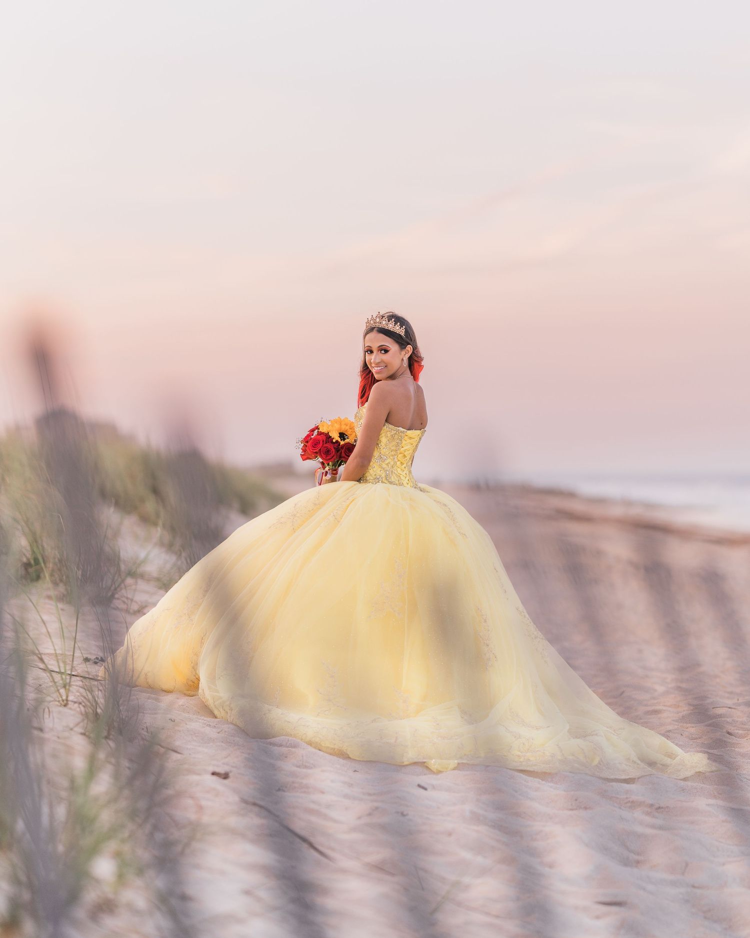 A person in a flowing yellow ballgown poses with a bouquet on a beach at sunset, creating a dreamy atmosphere.