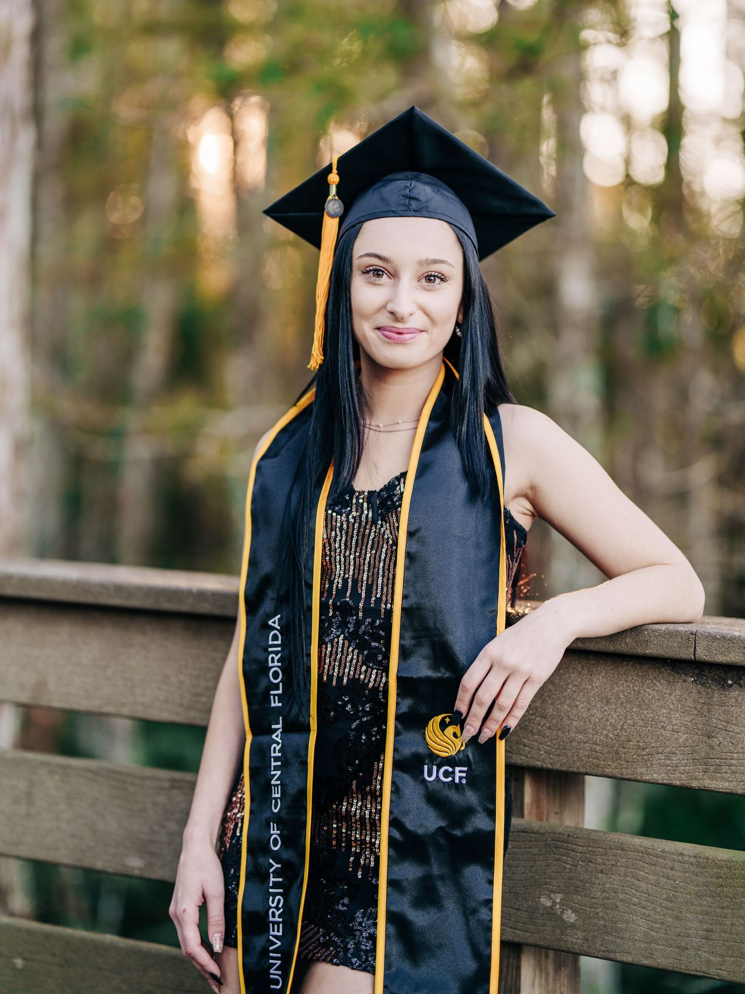 A graduate in black dress and cap wearing academic regalia with UCF sash poses by wooden railing in outdoor setting.