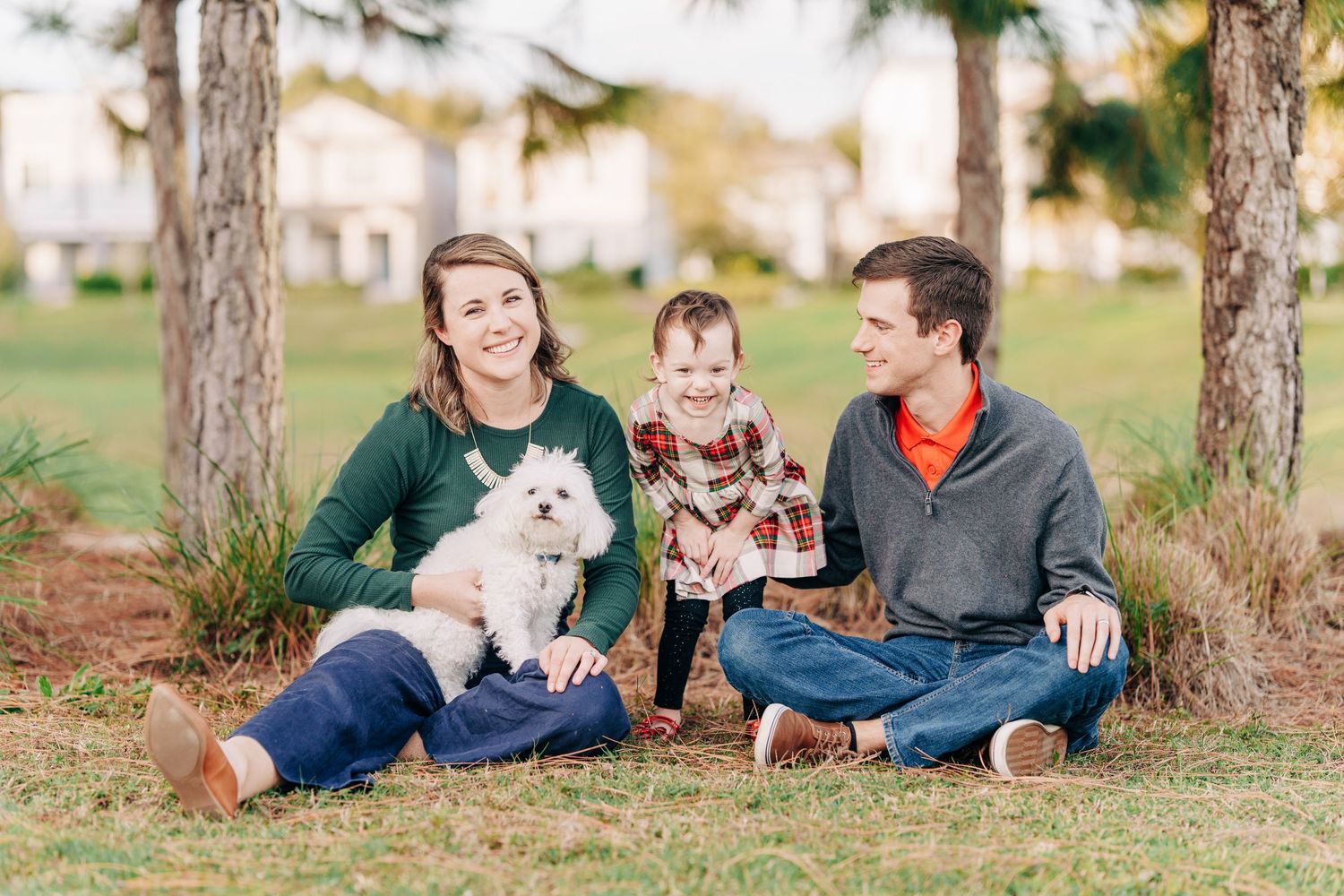 A family sits together outdoors on grass between trees with their small white dog.