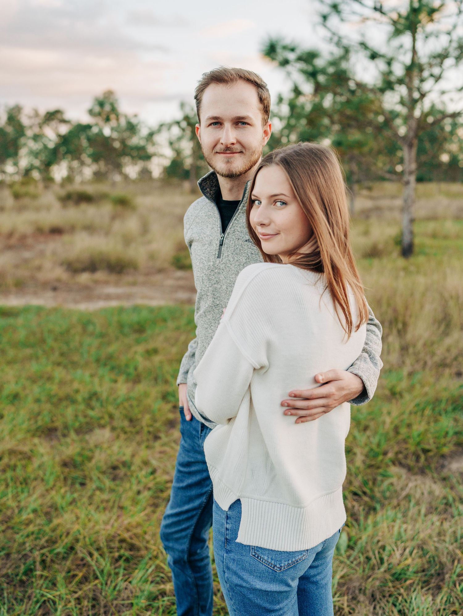 A couple in casual sweaters embrace while standing in a grassy field with trees at sunset.