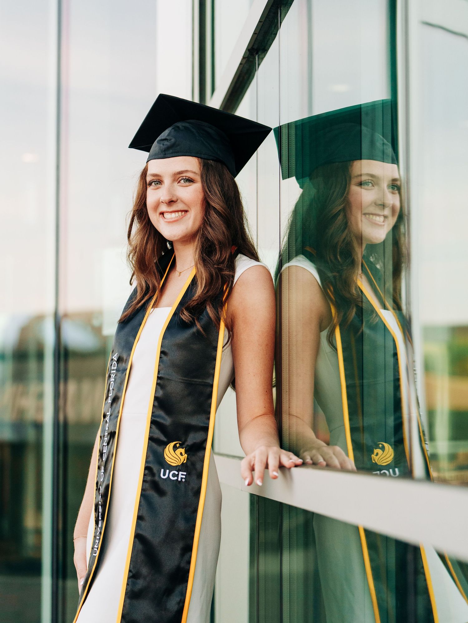 UCF Graduate in black cap and gown with gold sash poses by reflective glass window in outdoor setting.