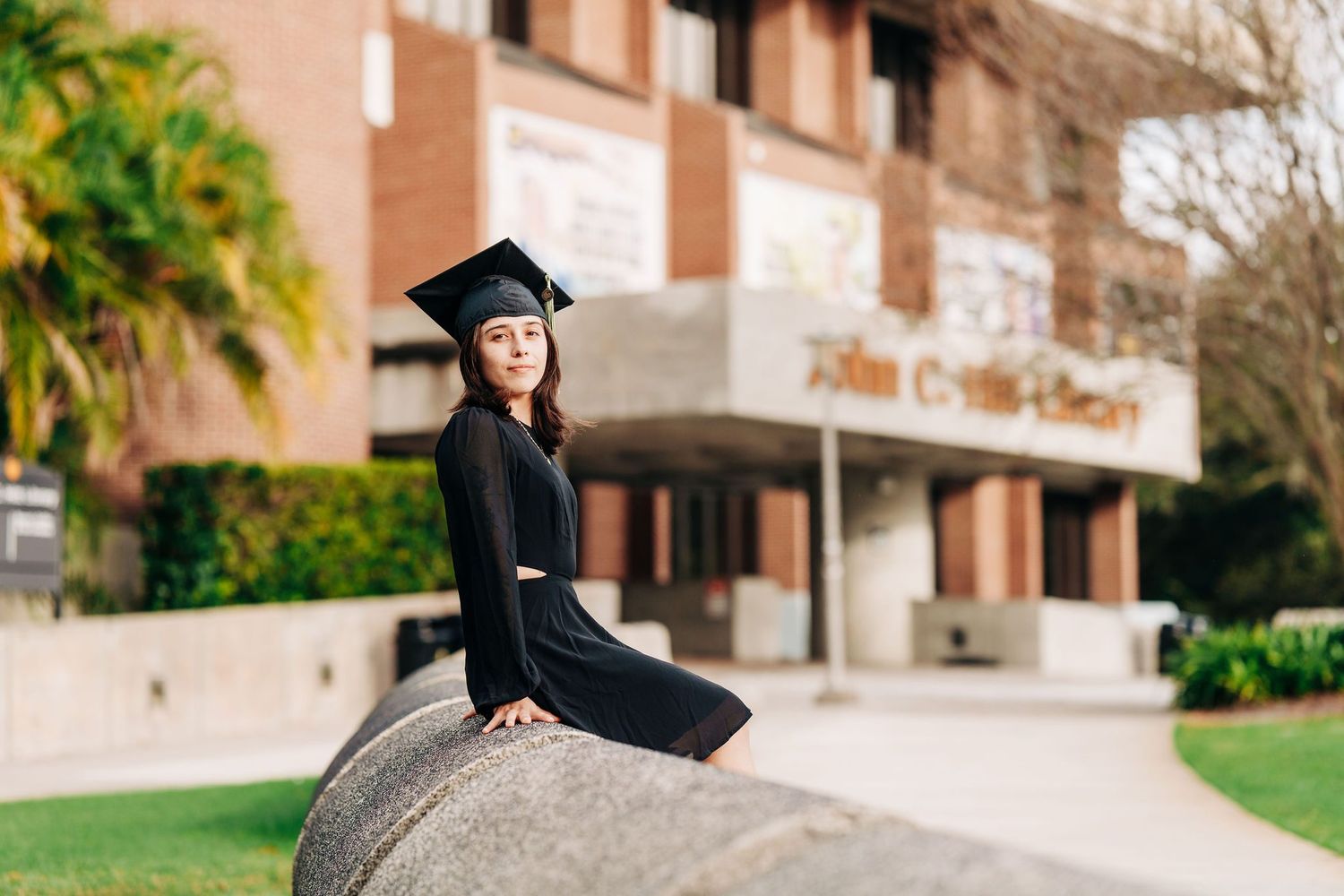 Woman in black graduation cap and gown sitting on stone sculpture outside university building.