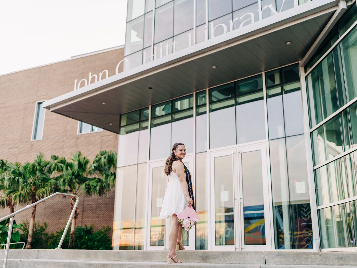 Young woman in pink dress poses on steps outside John C. Hitt Library building on a sunny day.