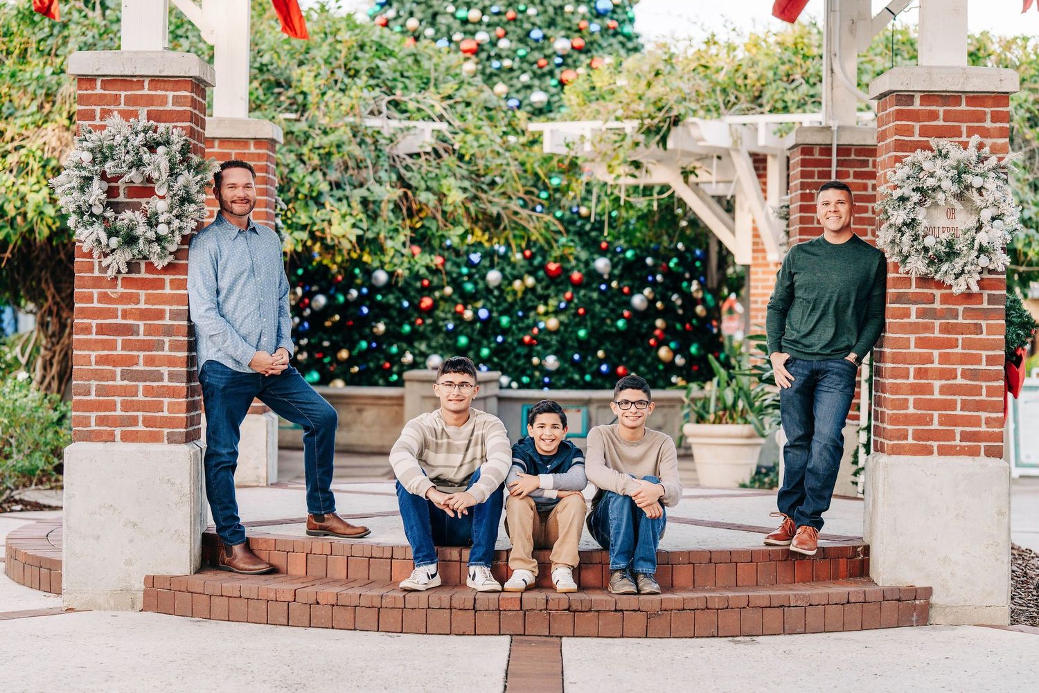 Family of five poses in front of a decorated Christmas tree between brick pillars for a holiday portrait.