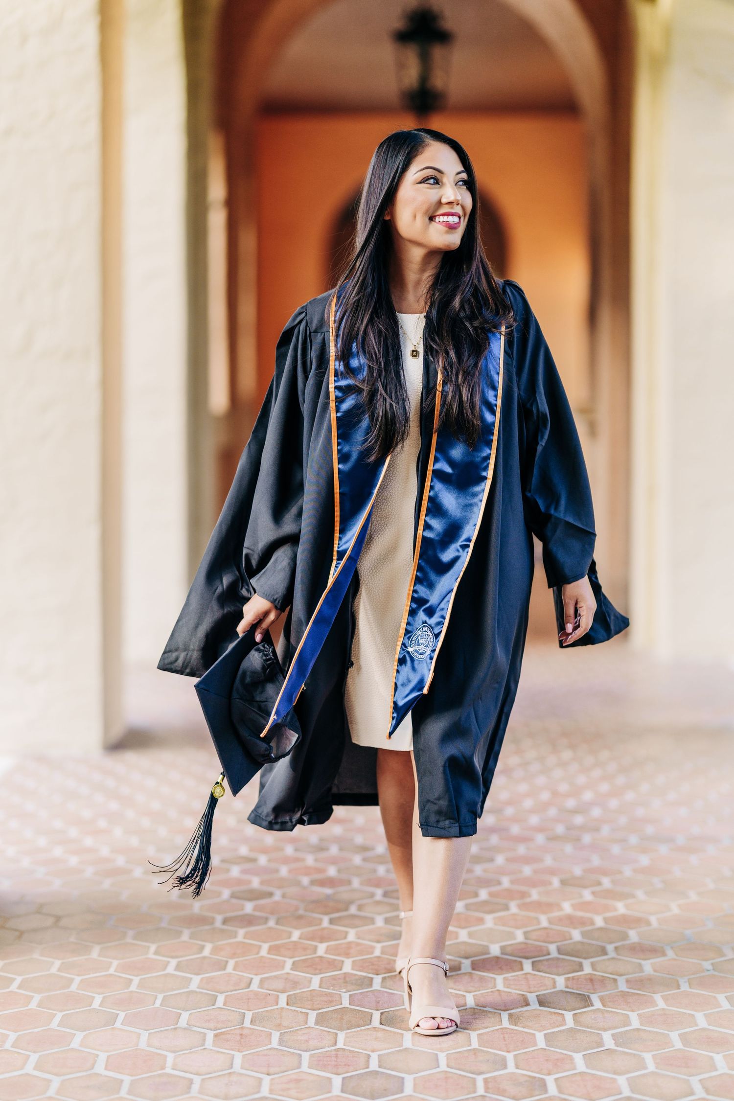 Graduate in cap and gown walks smiling on terracotta tile walkway beneath arched hallway during graduation photoshoot.