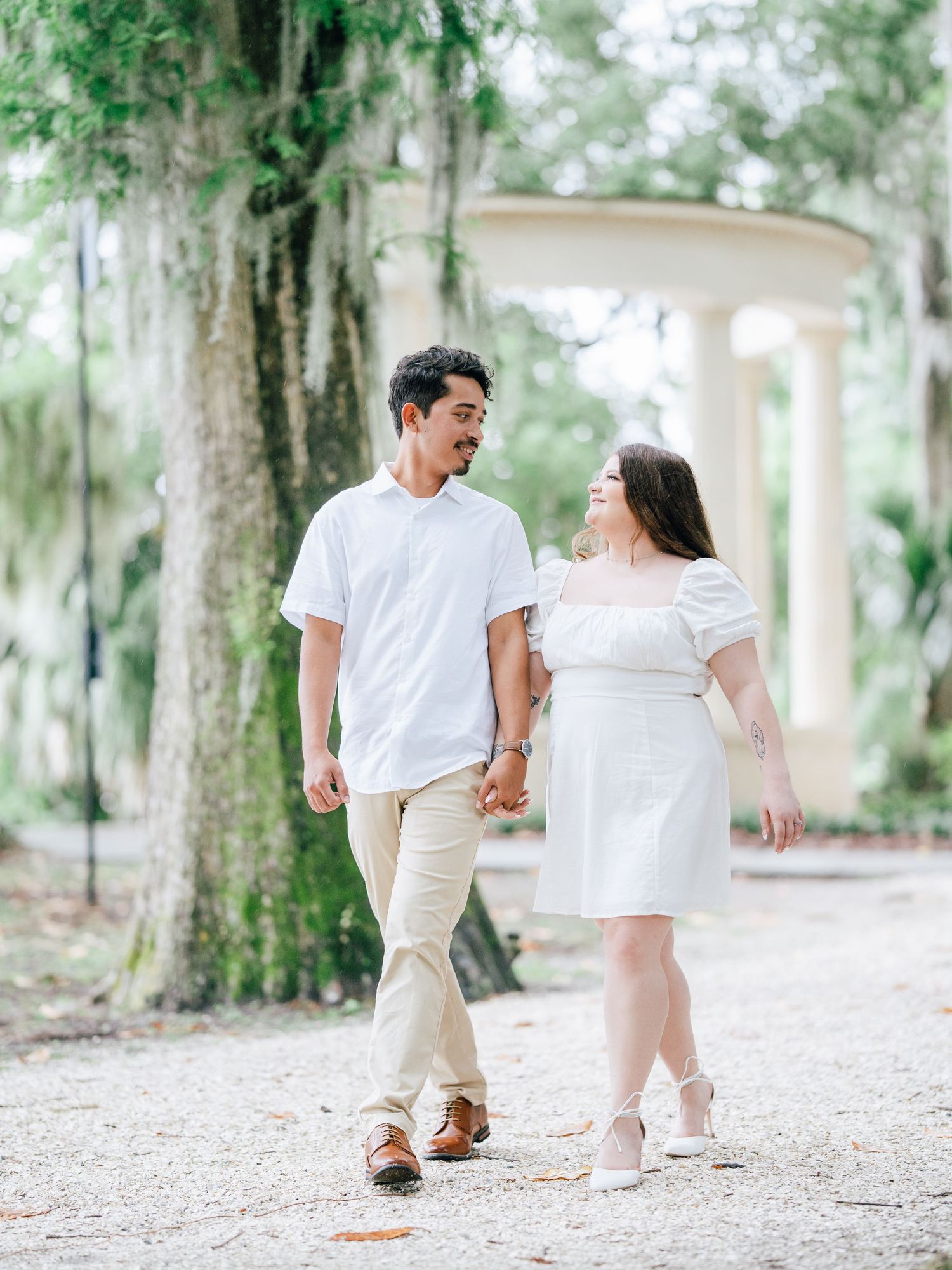 Couple in white outfits holding hands while walking on a garden path with Spanish moss hanging from trees.