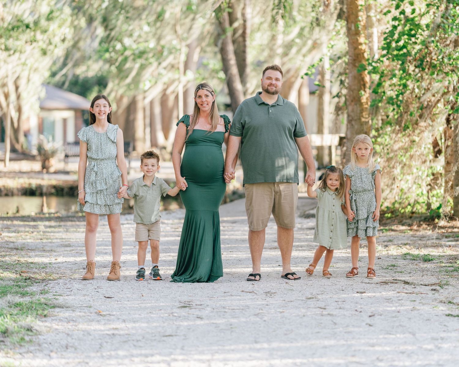 A family in matching casual outfits stands together on a tree-lined dirt path during a sunny day.