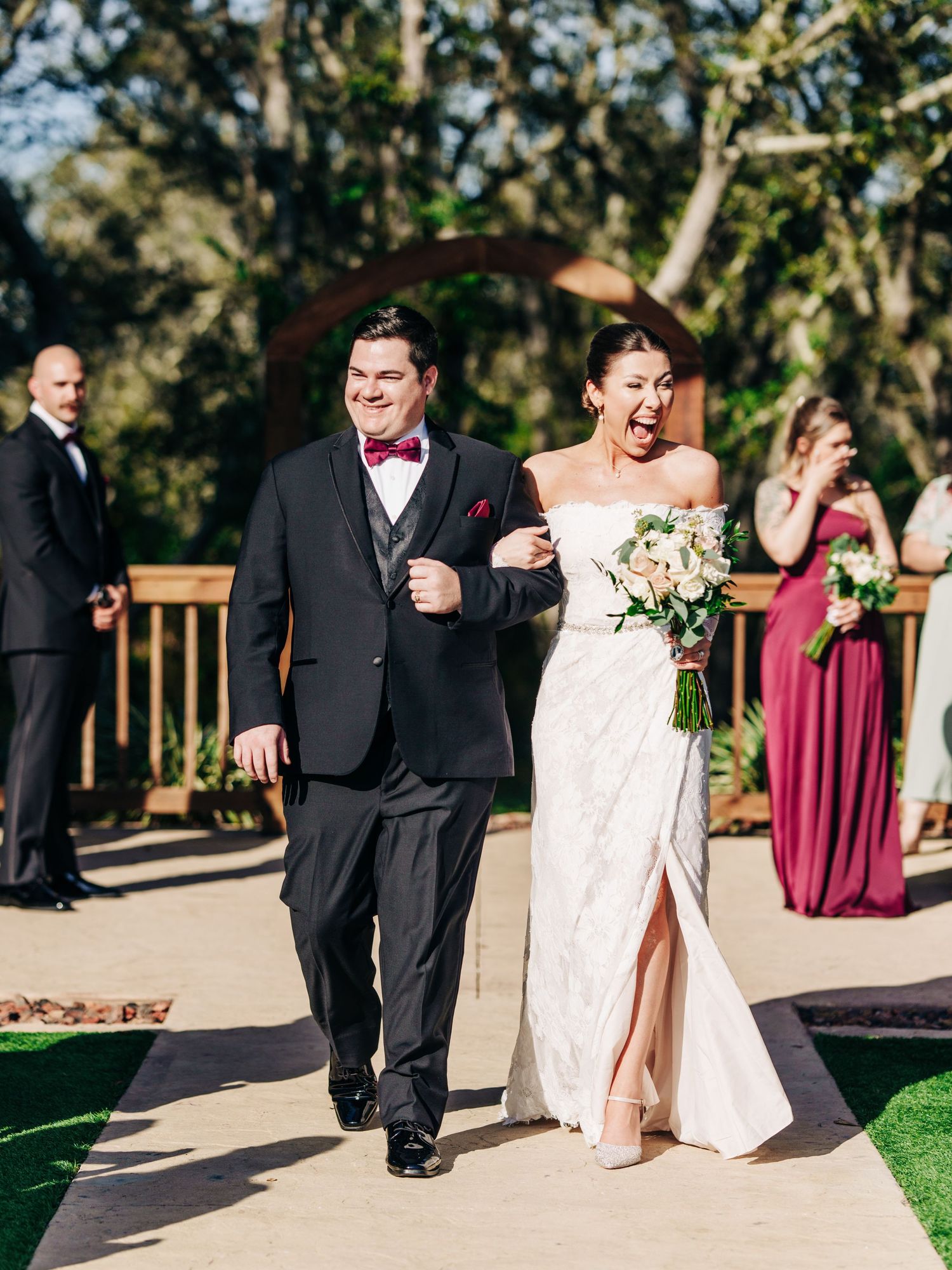 Wedding party members walk together on a sunny outdoor path during ceremony processional at elegant garden venue.