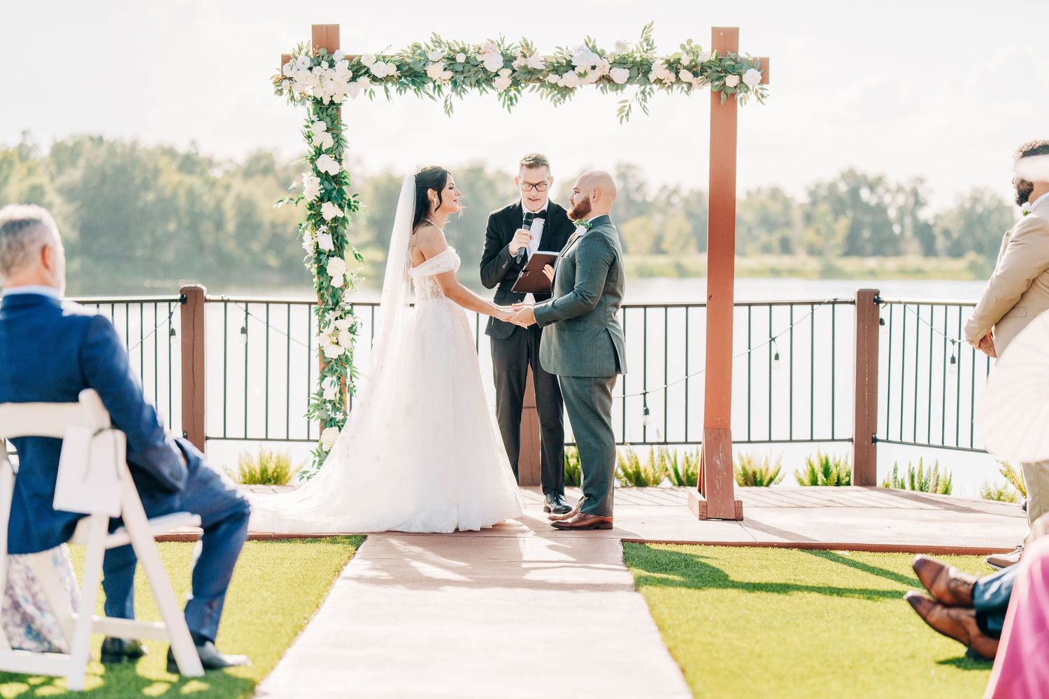 Outdoor wedding ceremony on a deck with floral arch, bride and groom exchanging vows with officiant, lake in background.