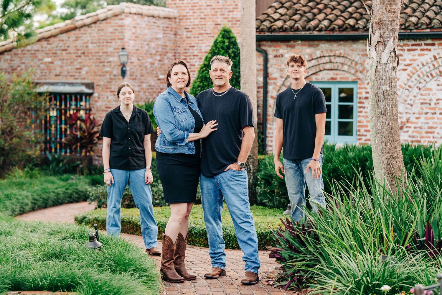 Family of four posing outdoors in front of a brick home surrounded by lush greenery.