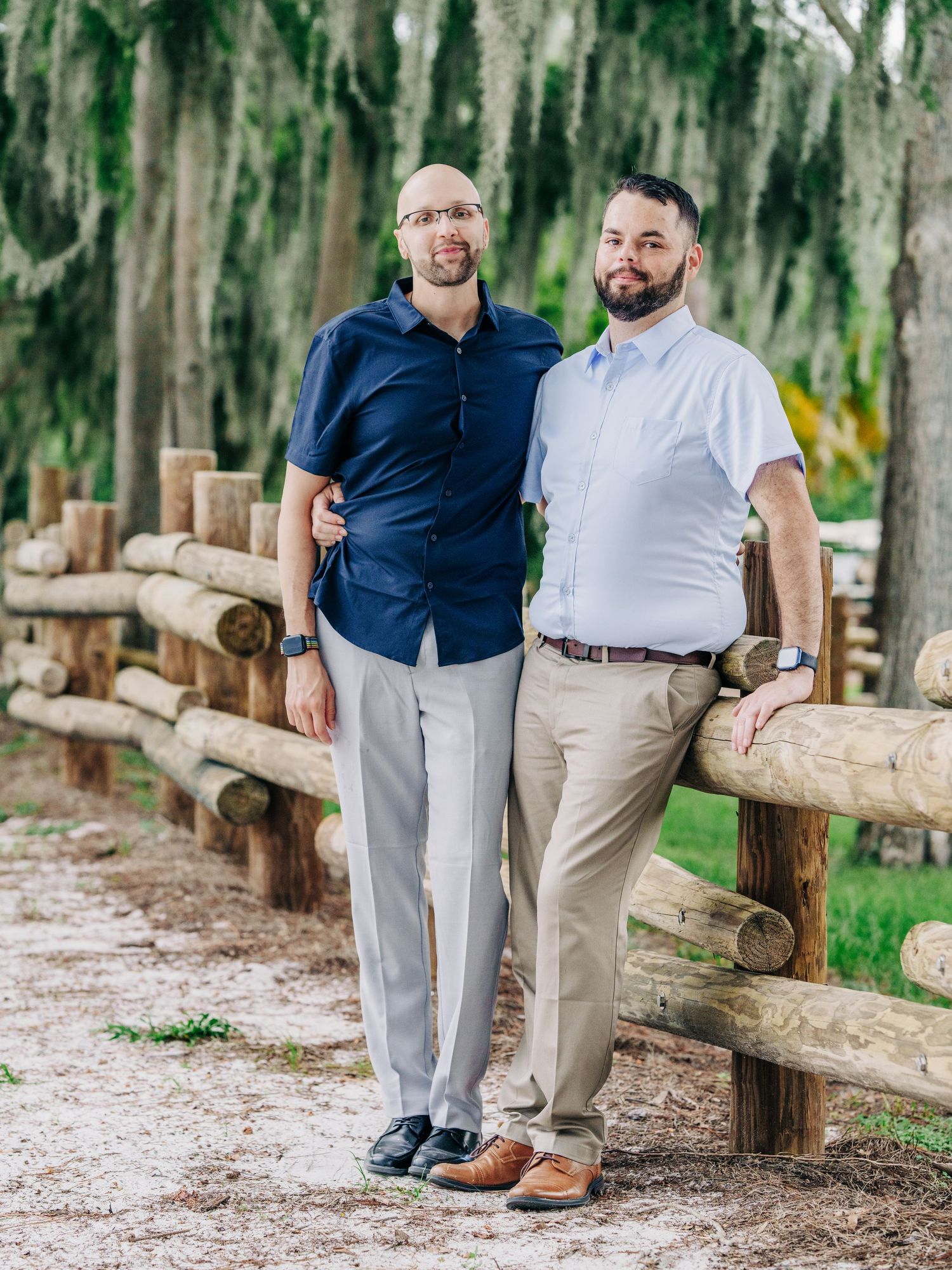 A same-sex couple pose together by a rustic wooden fence in a lush outdoor setting with Spanish moss.