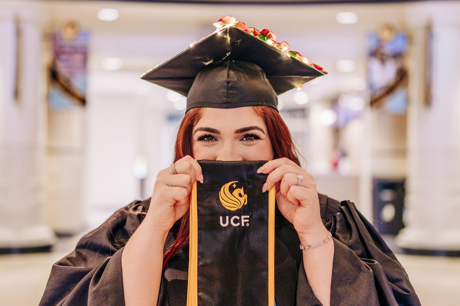 UCF graduate in black cap and gown holding diploma cover over face at University of Central Florida ceremony.
