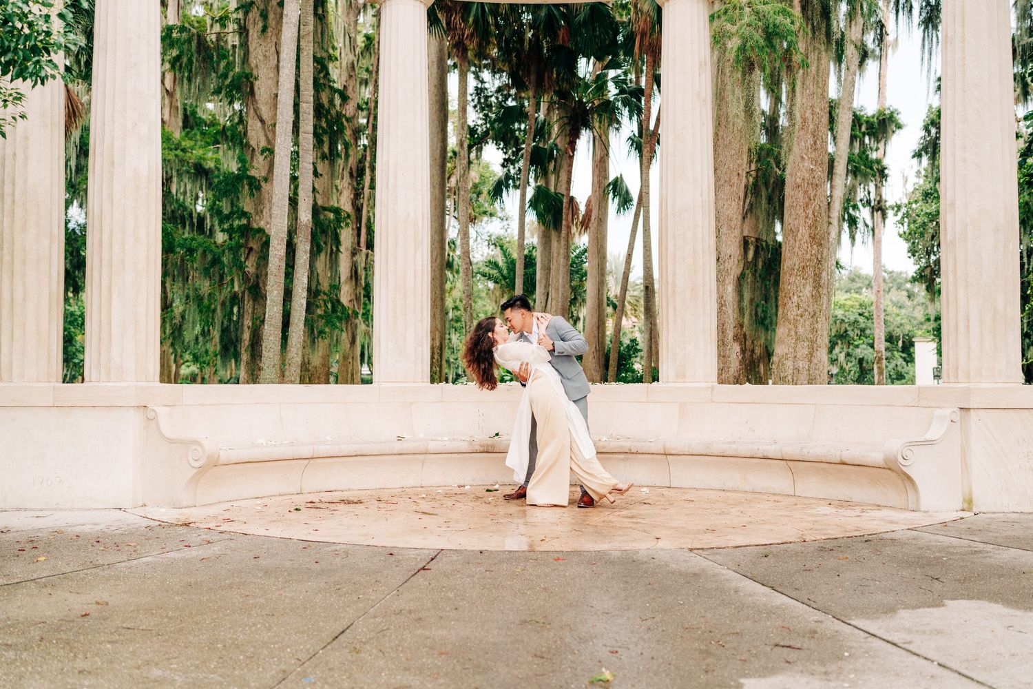 Couple sharing a romantic dip kiss under grand white columns surrounded by lush tropical trees at an outdoor wedding venue.