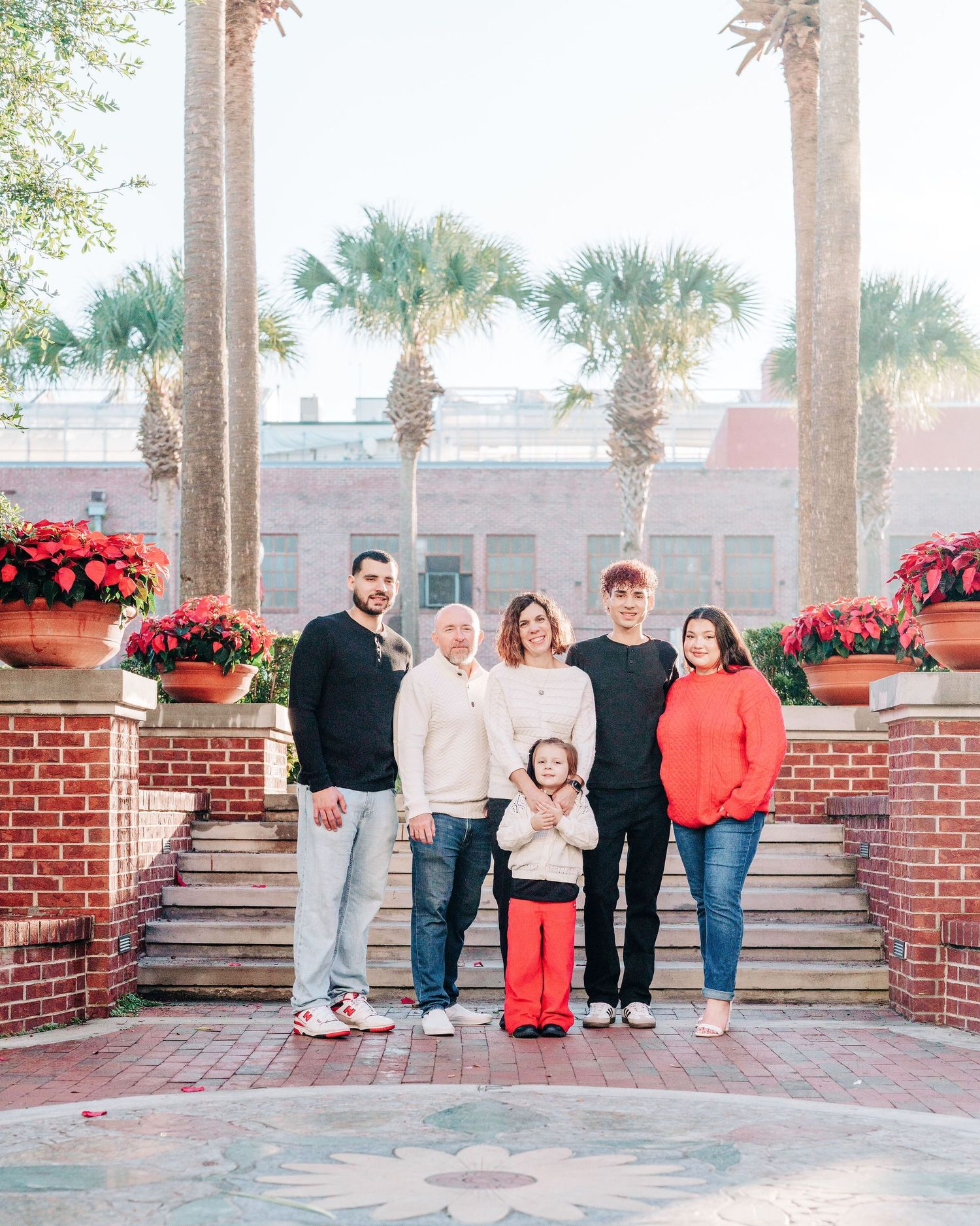 Family of six posing outdoors near brick columns with red poinsettias and palm trees in festive holiday setting.