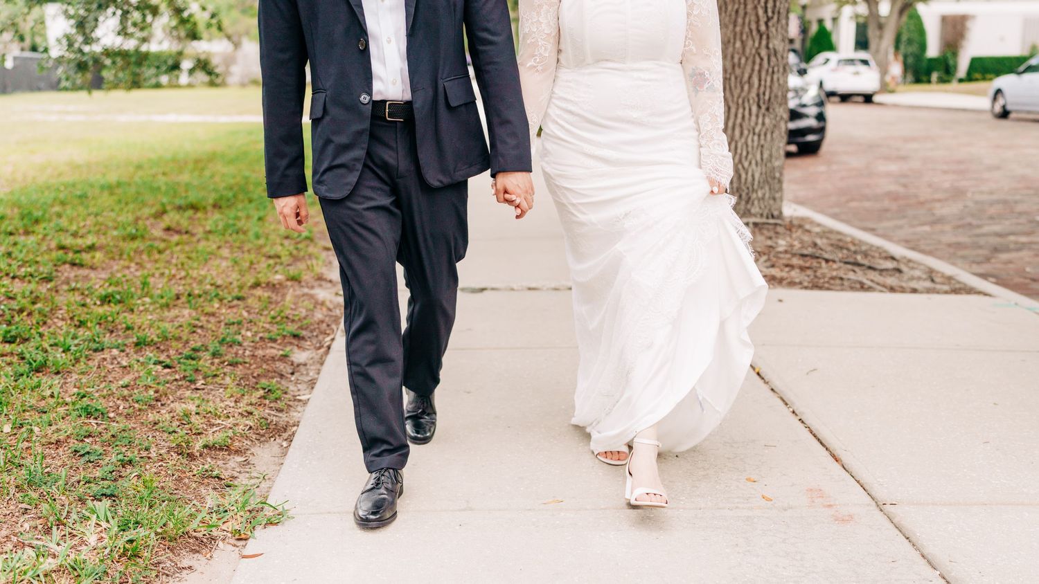 Bride and groom walking hand in hand on a sidewalk, wearing wedding attire on their special day.