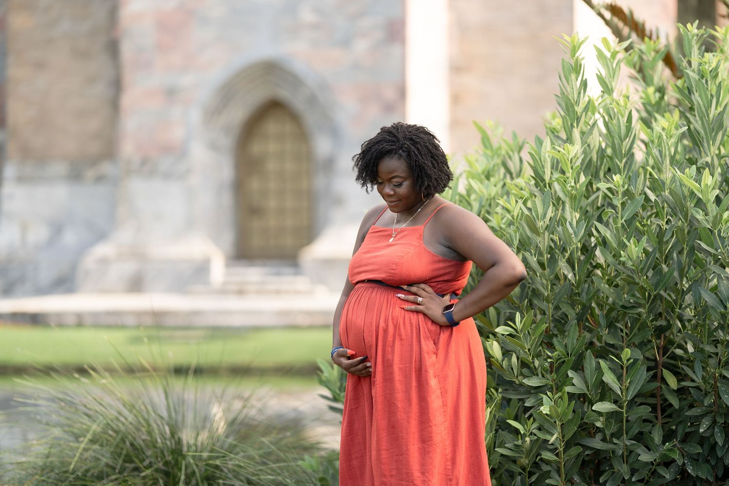 A pregnant woman in a flowing coral dress stands beside green foliage at Bok Tower Gardens.