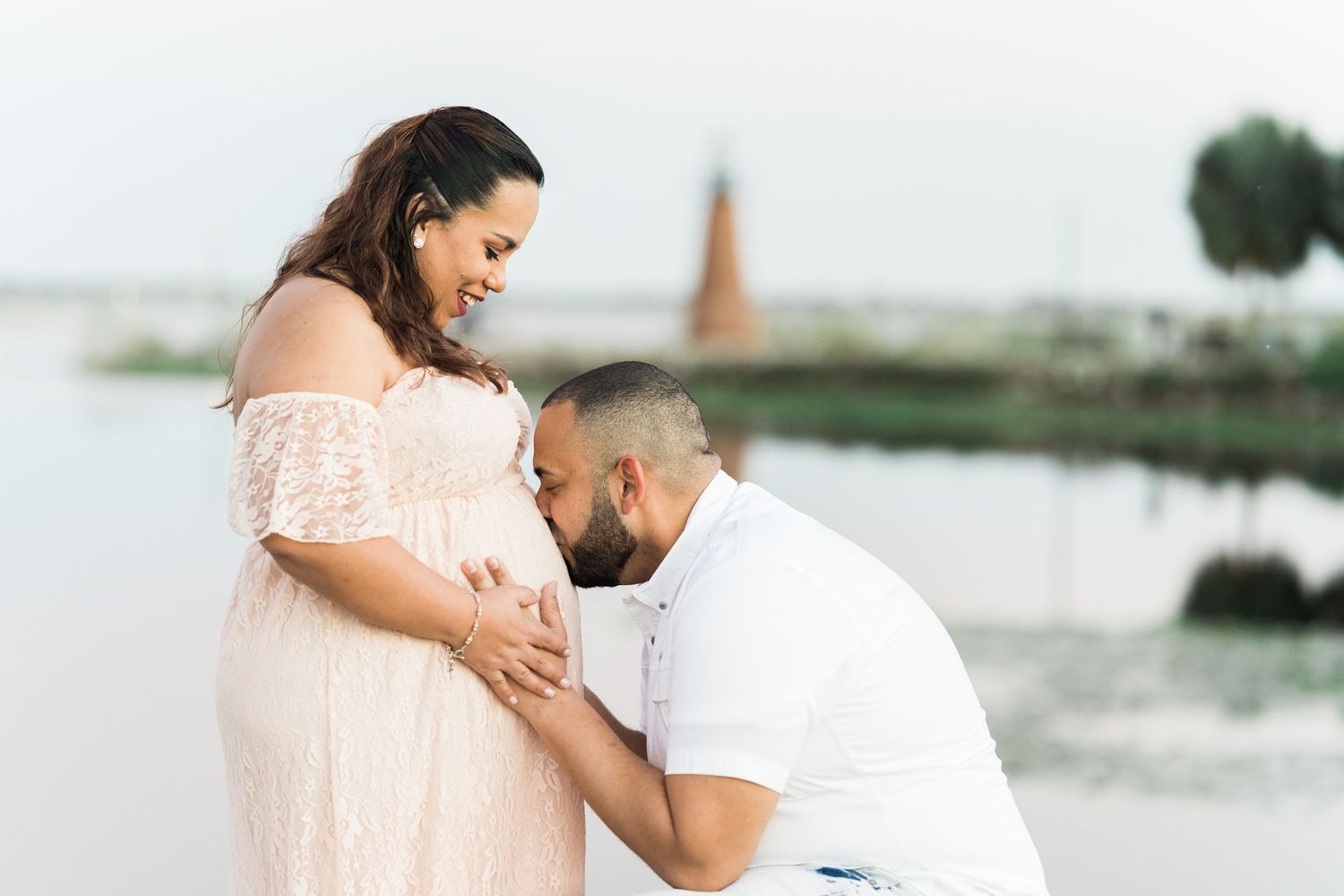 A couple shares a tender moment by the water, with the person in a white dress while their partner kisses their belly.