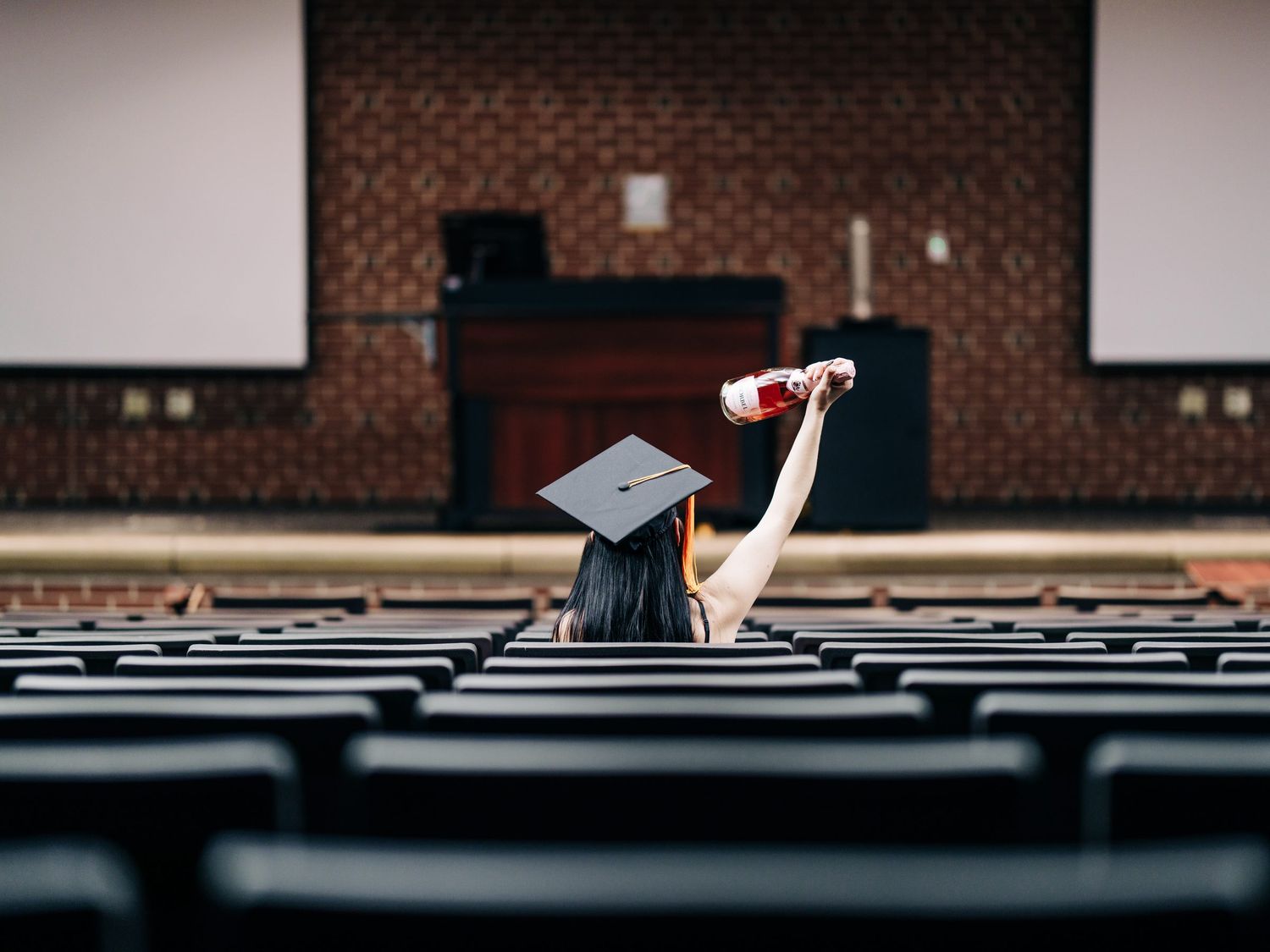 Graduate in cap raises diploma triumphantly in empty auditorium after graduation ceremony.