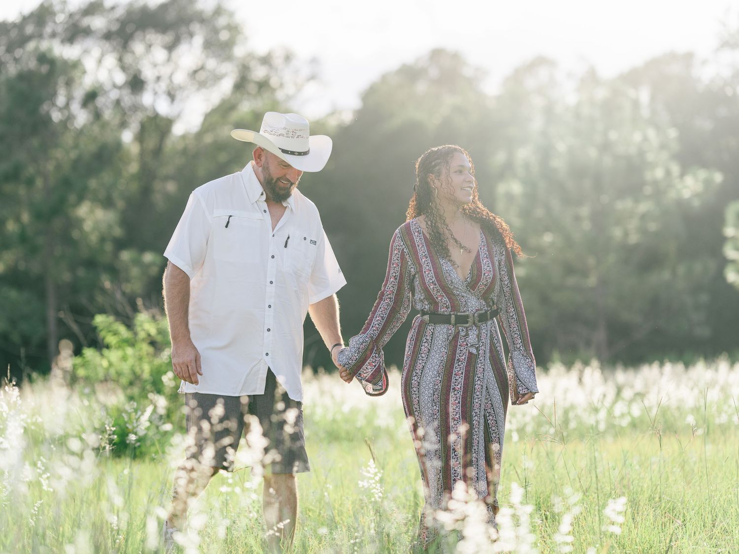 Two people holding hands while walking through a sunlit meadow with wildflowers on a bright summer day.