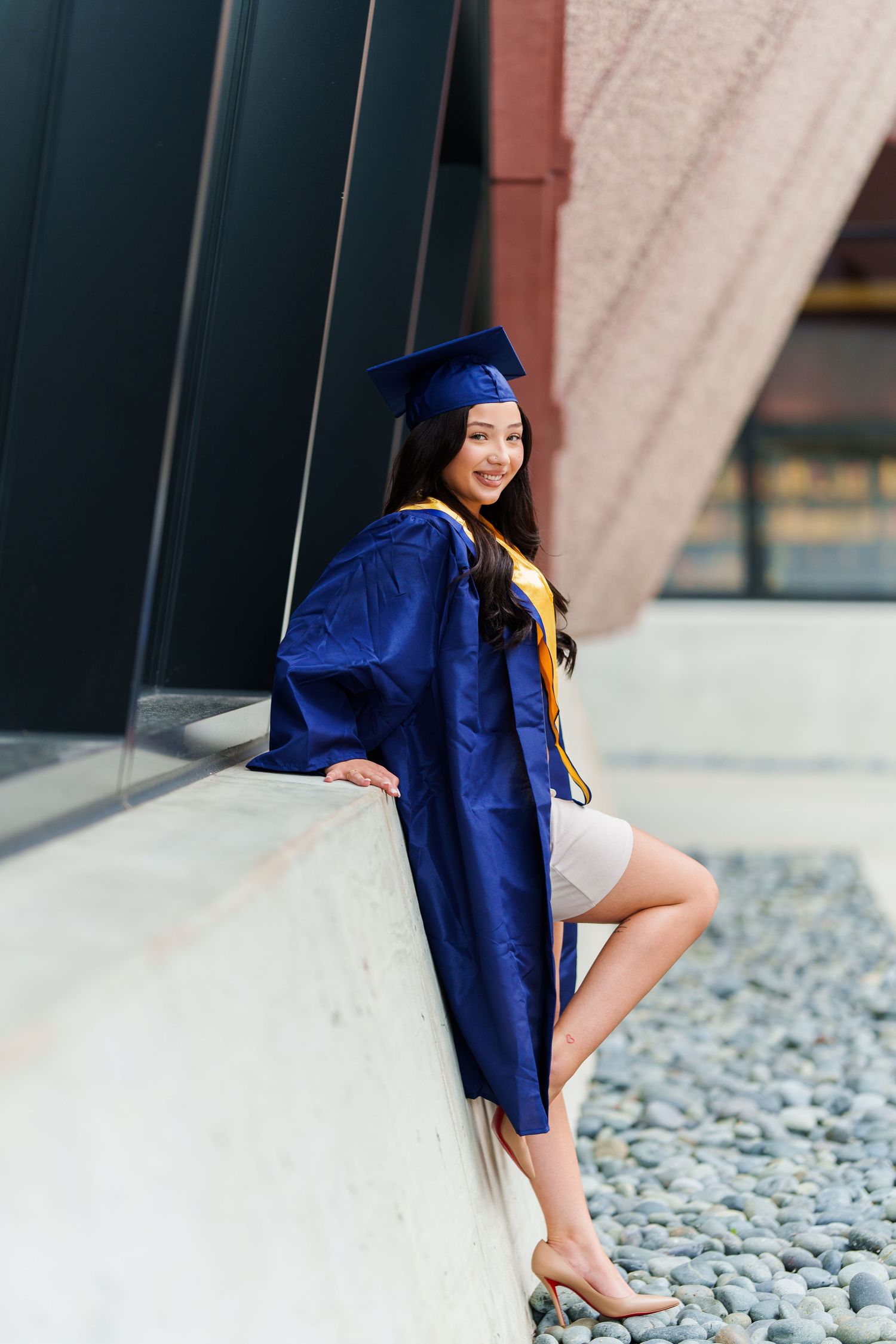 Graduate in blue cap and gown poses on concrete ledge during outdoor graduation photo session.