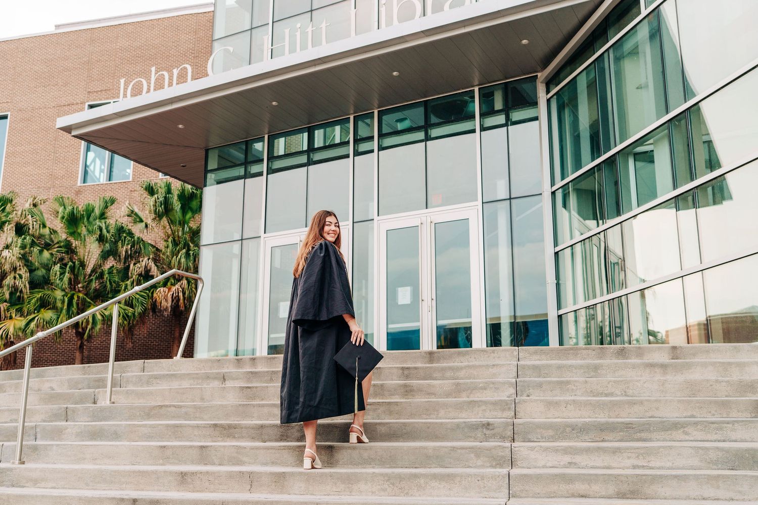 A smiling graduate in black cap and gown poses on university steps holding her mortarboard outside a modern glass building.