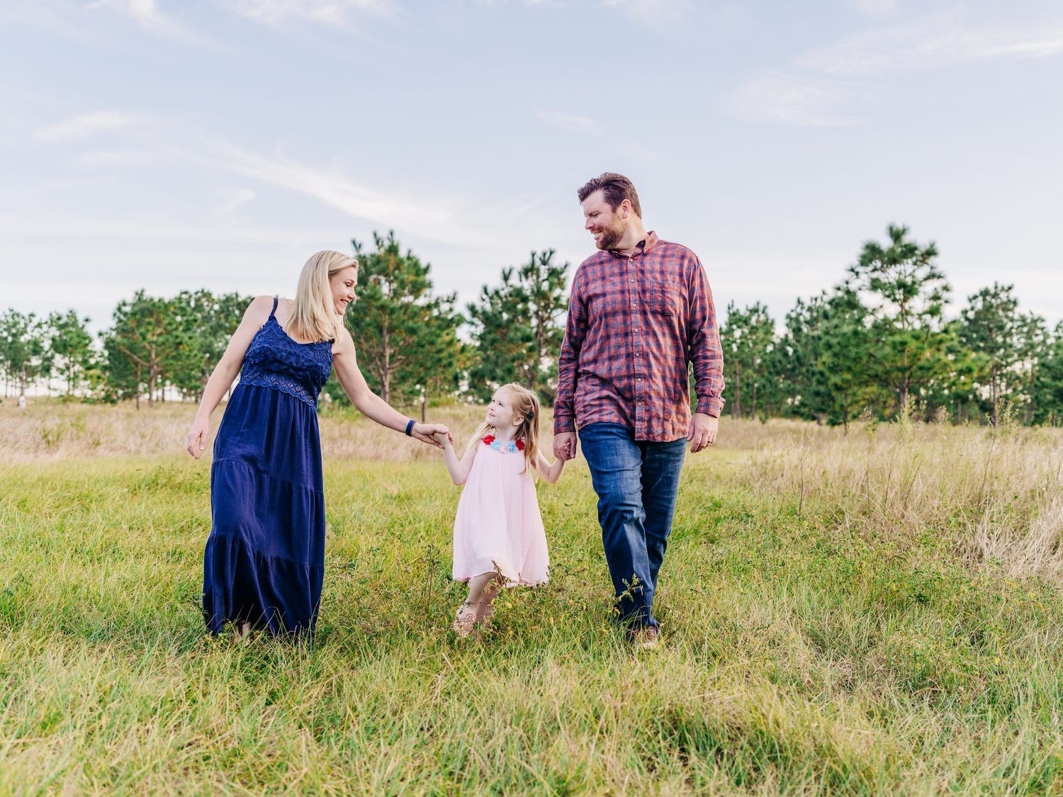 A happy family of three walks through a green meadow, parents holding their young daughter's hands under a blue sky.