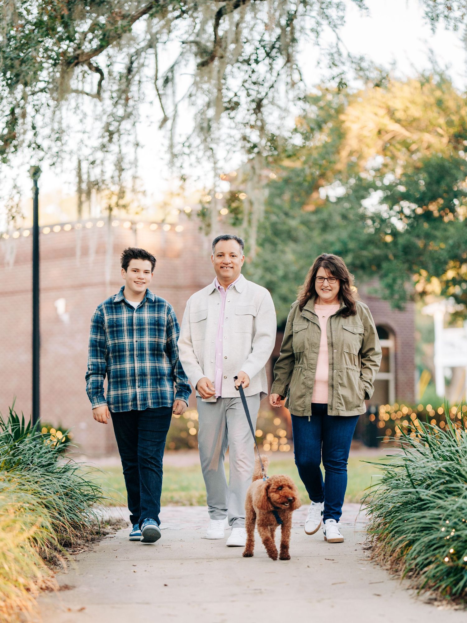 Three people walking an adorable brown goldendoodle dog on a pathway lined with plants during golden hour.