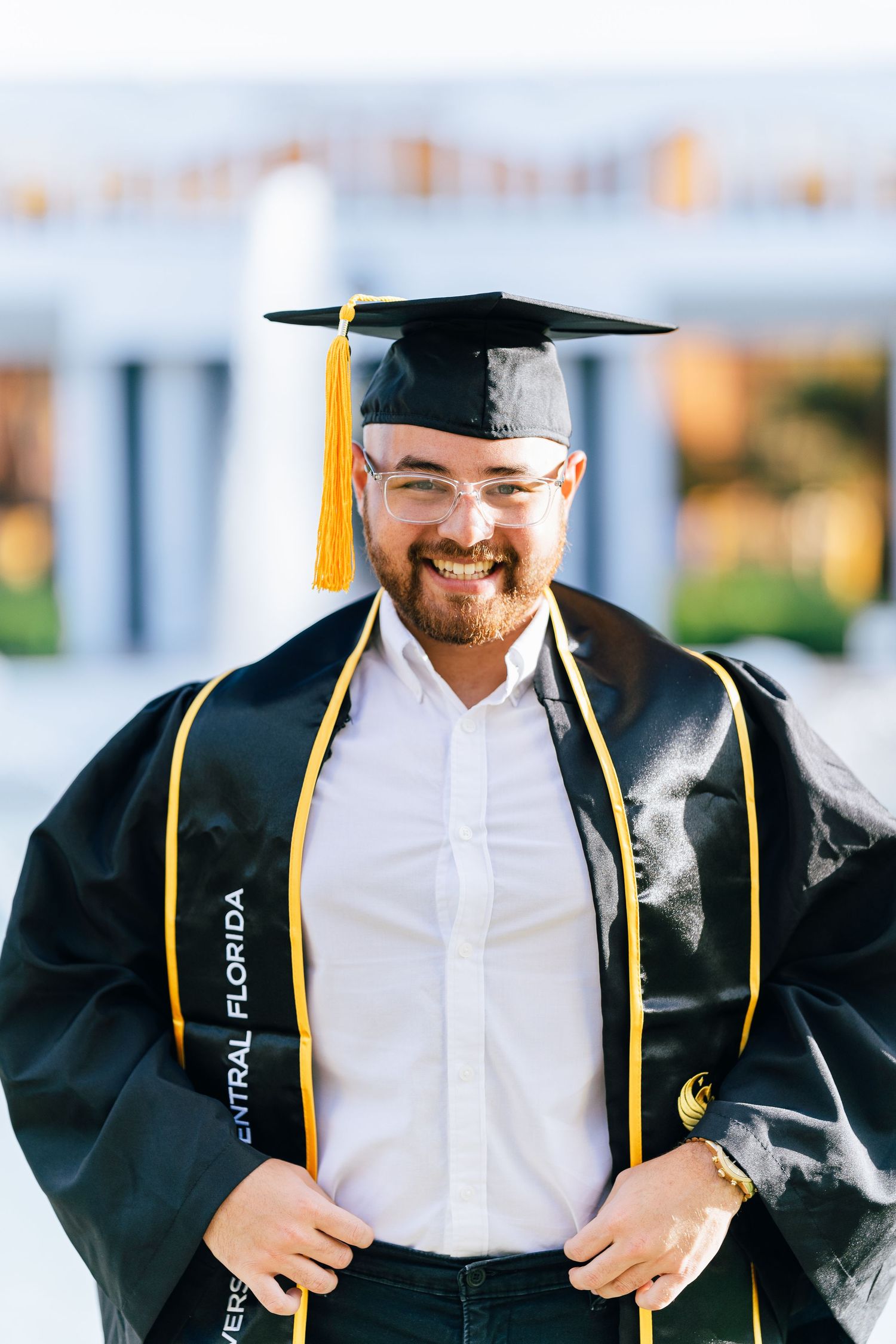 Graduate wearing black cap and gown with gold honor cords stands smiling outdoors on graduation day.