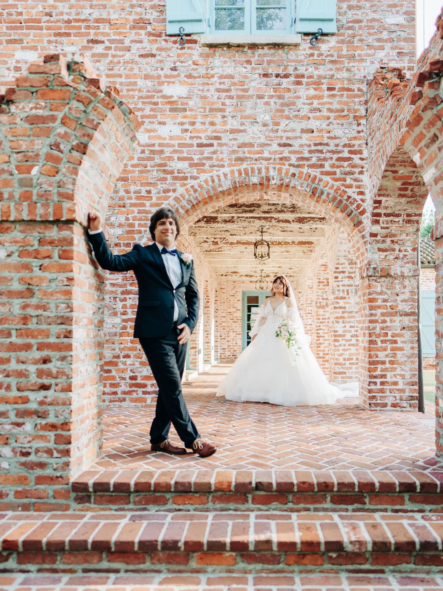 A bride and groom pose beneath brick archways at a historic venue with teal shutters in the background.