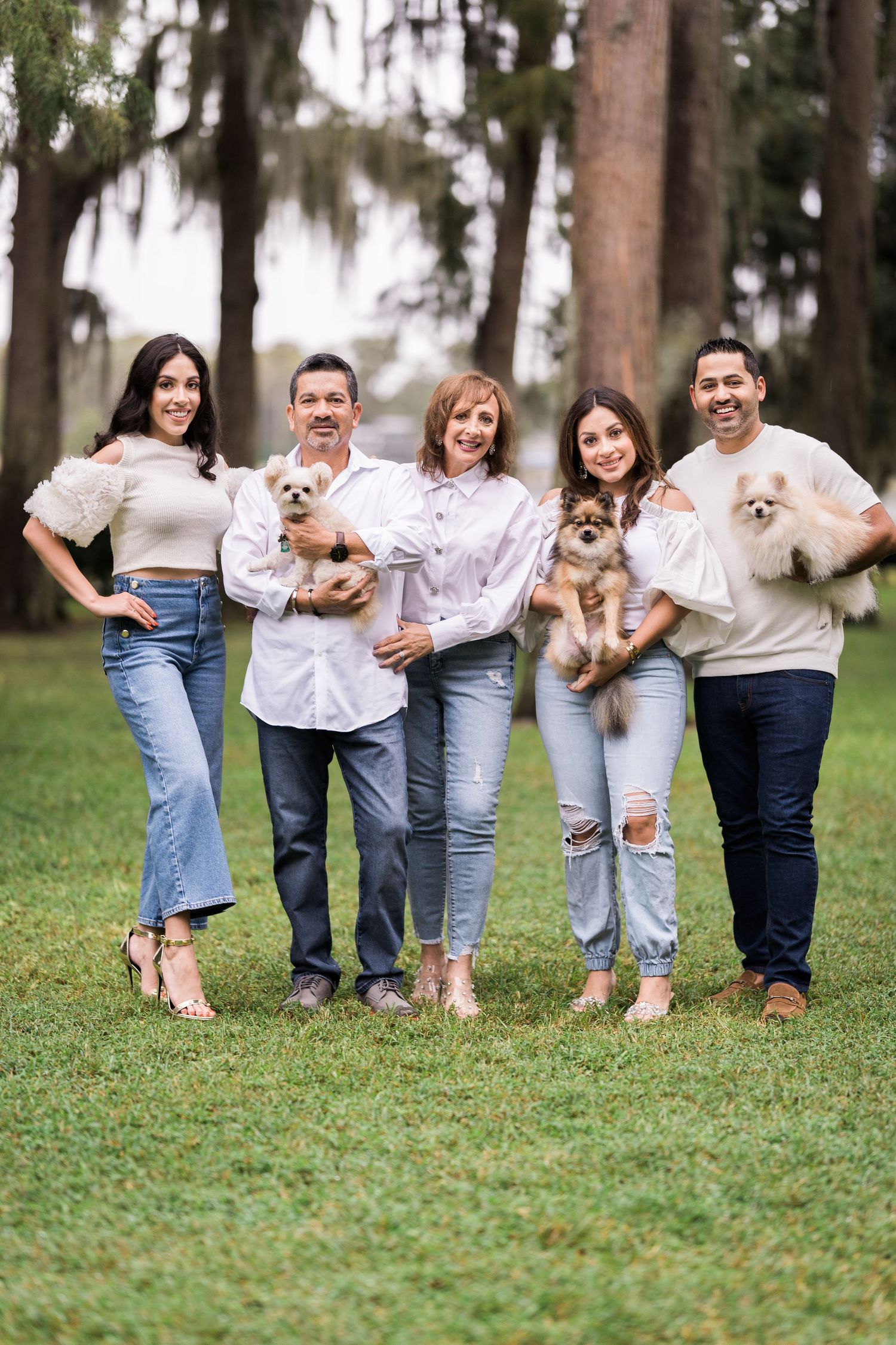A group stands together on grassy lawn while holding small dogs, wearing casual white and denim outfits.