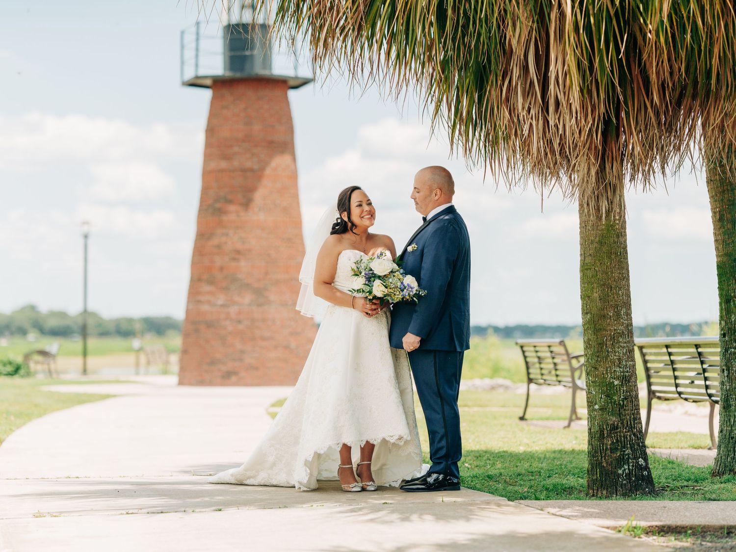 A wedding couple stands together under a palm tree near a brick lighthouse on a sunny day holding a bouquet.