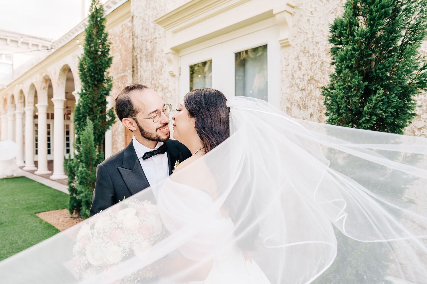 Bride and groom share a romantic kiss outdoors, with her veil flowing in the wind near an elegant stone building.