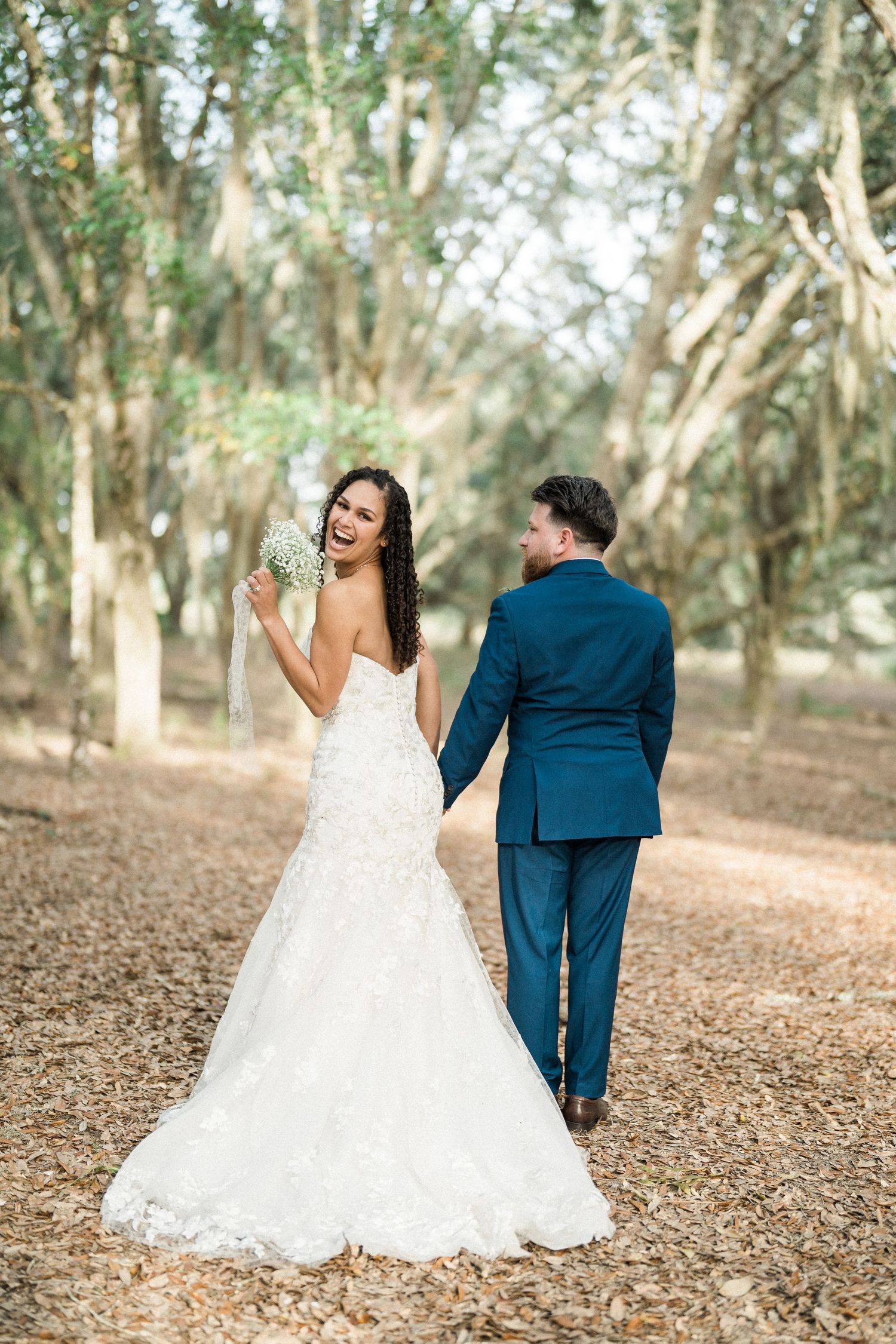 A bride in a white lace dress and groom in navy suit walk together on a tree-lined path during their outdoor wedding.