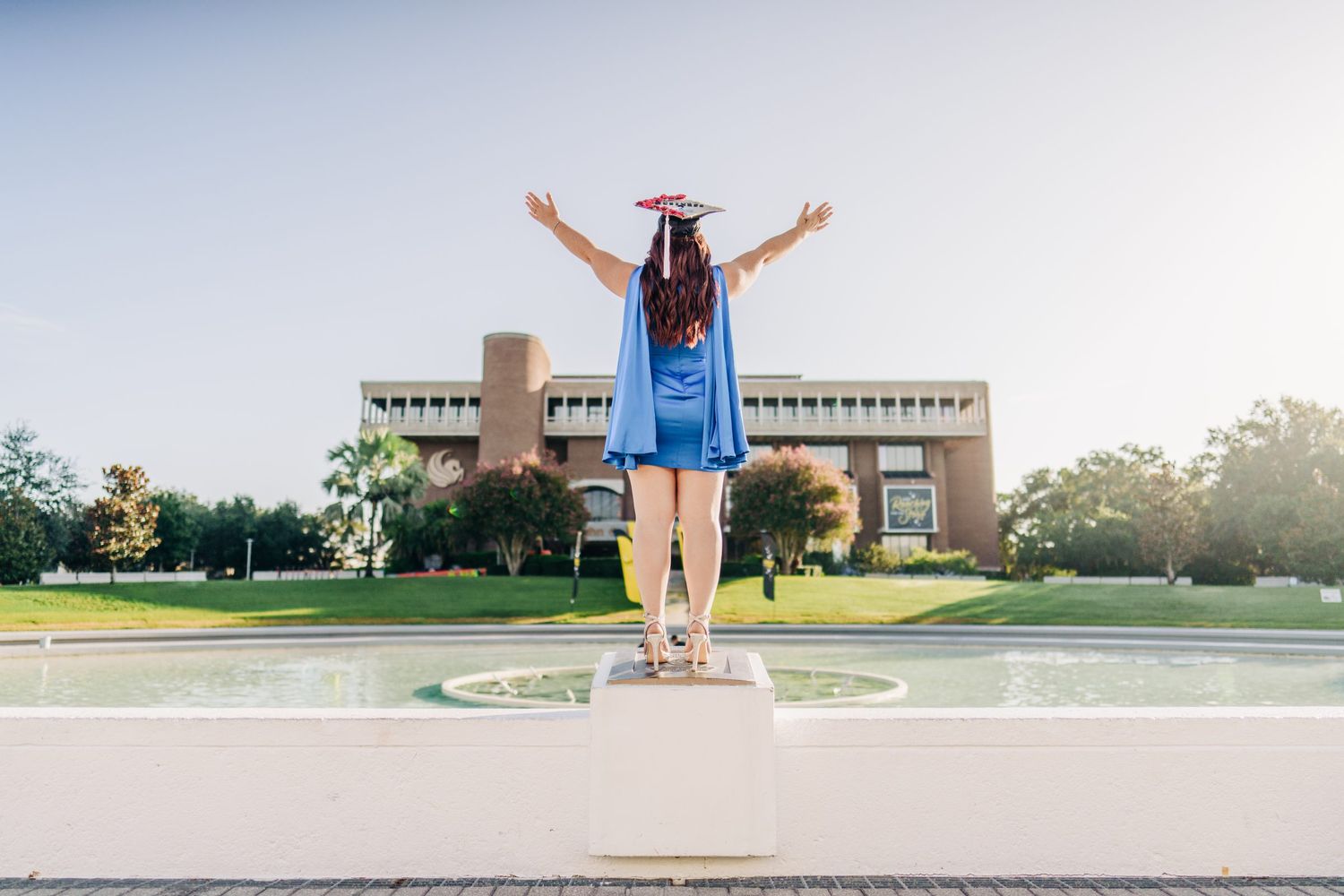 Graduate in blue gown and cap stands triumphantly with raised arms before a university fountain.