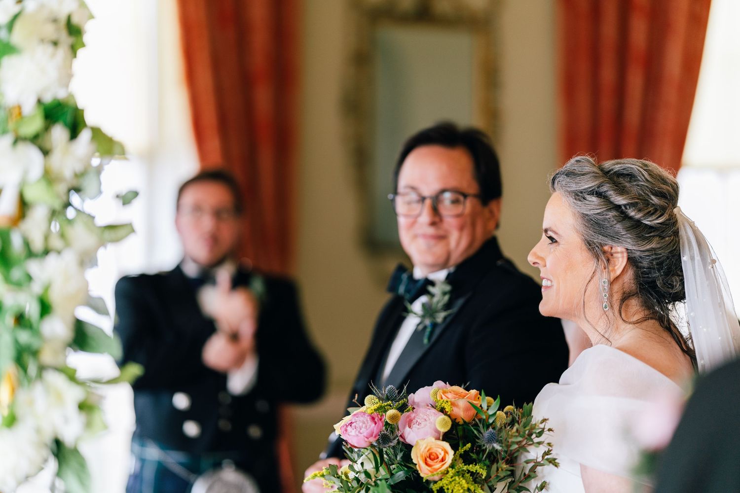 A bride holding a colorful bouquet of pink and peach flowers stands inside an elegant room with red curtains.