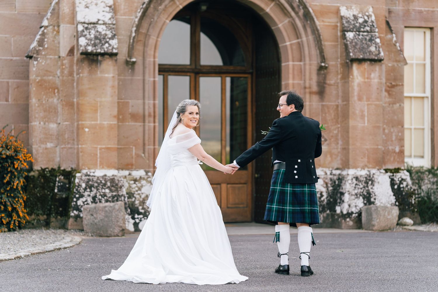 A newlywed couple in traditional Scottish wedding attire holds hands while standing at a stone church entrance.
