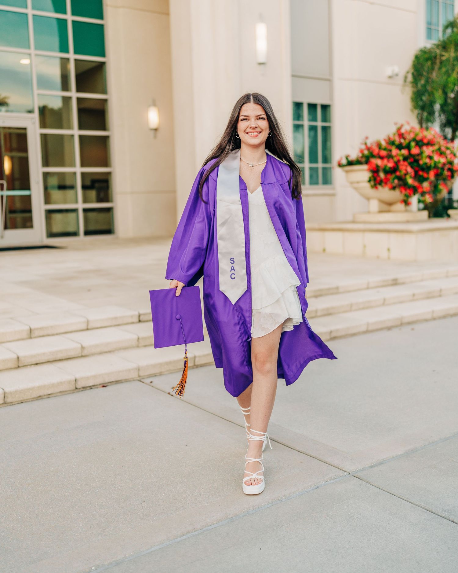 Graduate in bright purple academic regalia walks with matching diploma holder on steps of campus building.