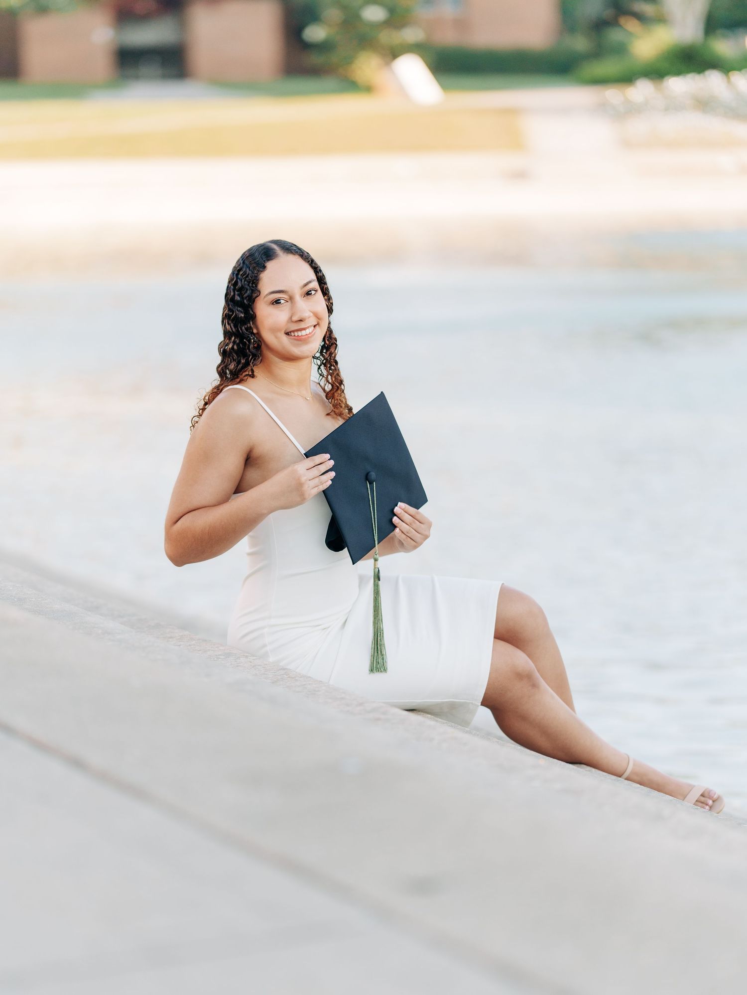 Graduate in a white dress holding a black diploma folder poses on a sunny beach.