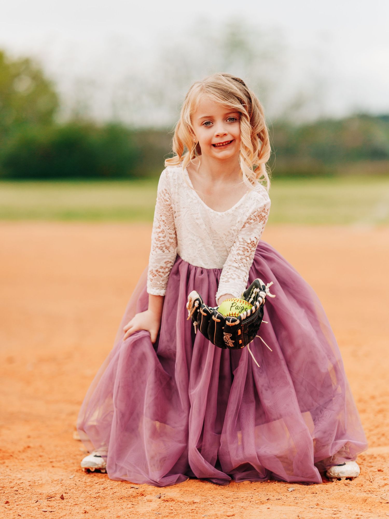 A young girl stands on an orange dirt path wearing a lace top and mauve tulle skirt while carrying a small basket.