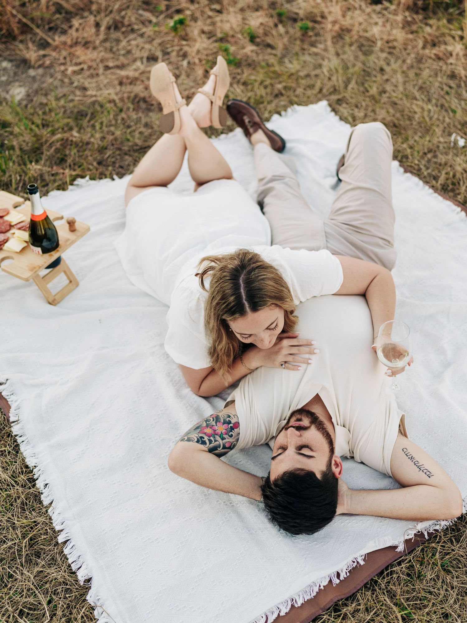 Couple relaxing together on white blanket during romantic outdoor picnic with wine and snacks.