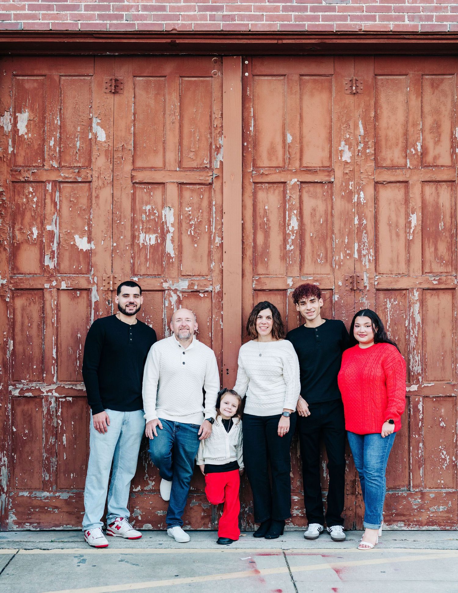 Family of six posing together in front of rustic wooden doors, wearing casual outfits in red, white, and black.