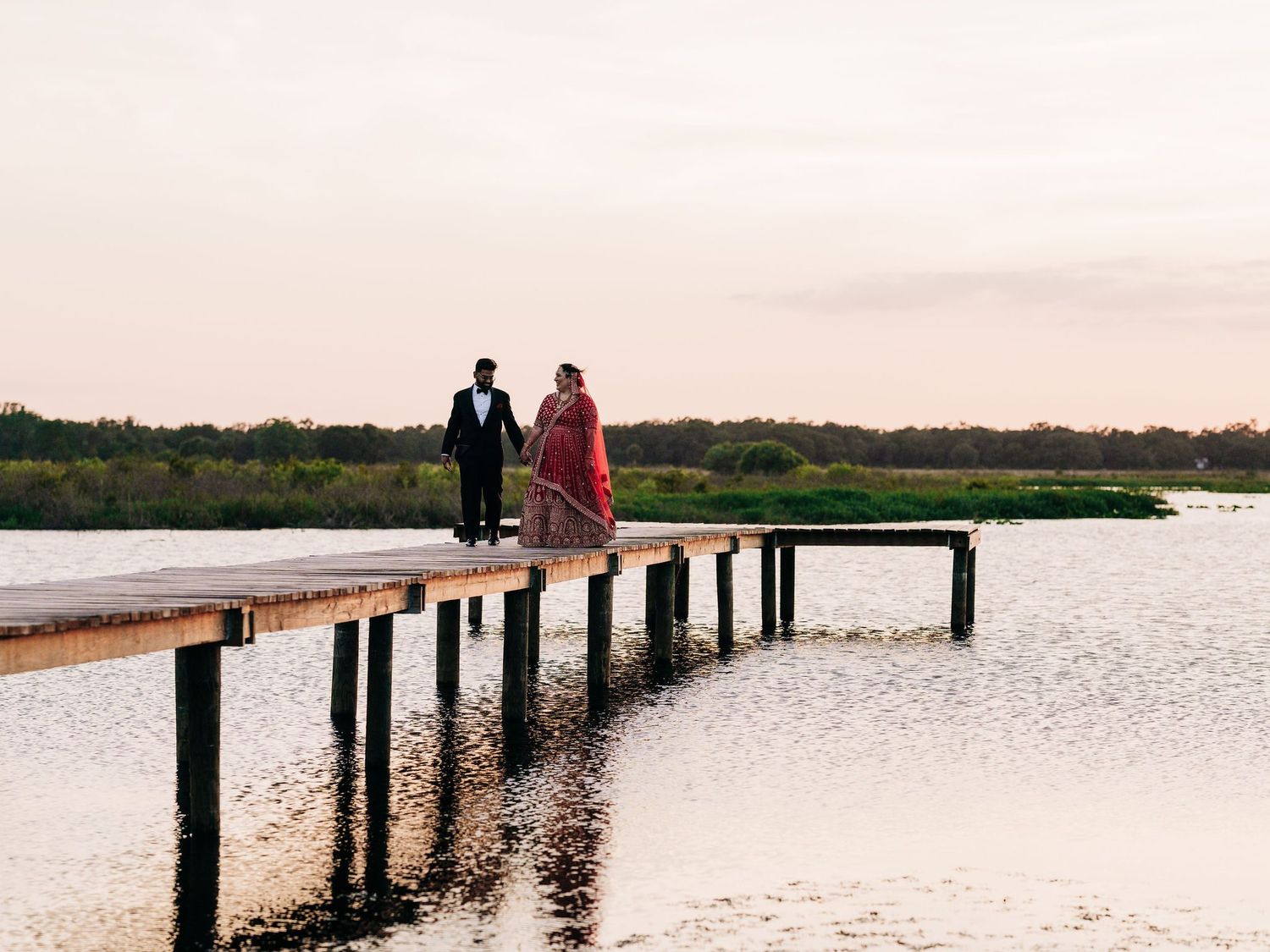 Couple in formal attire standing on a wooden dock over calm water at sunset with lush greenery in background.