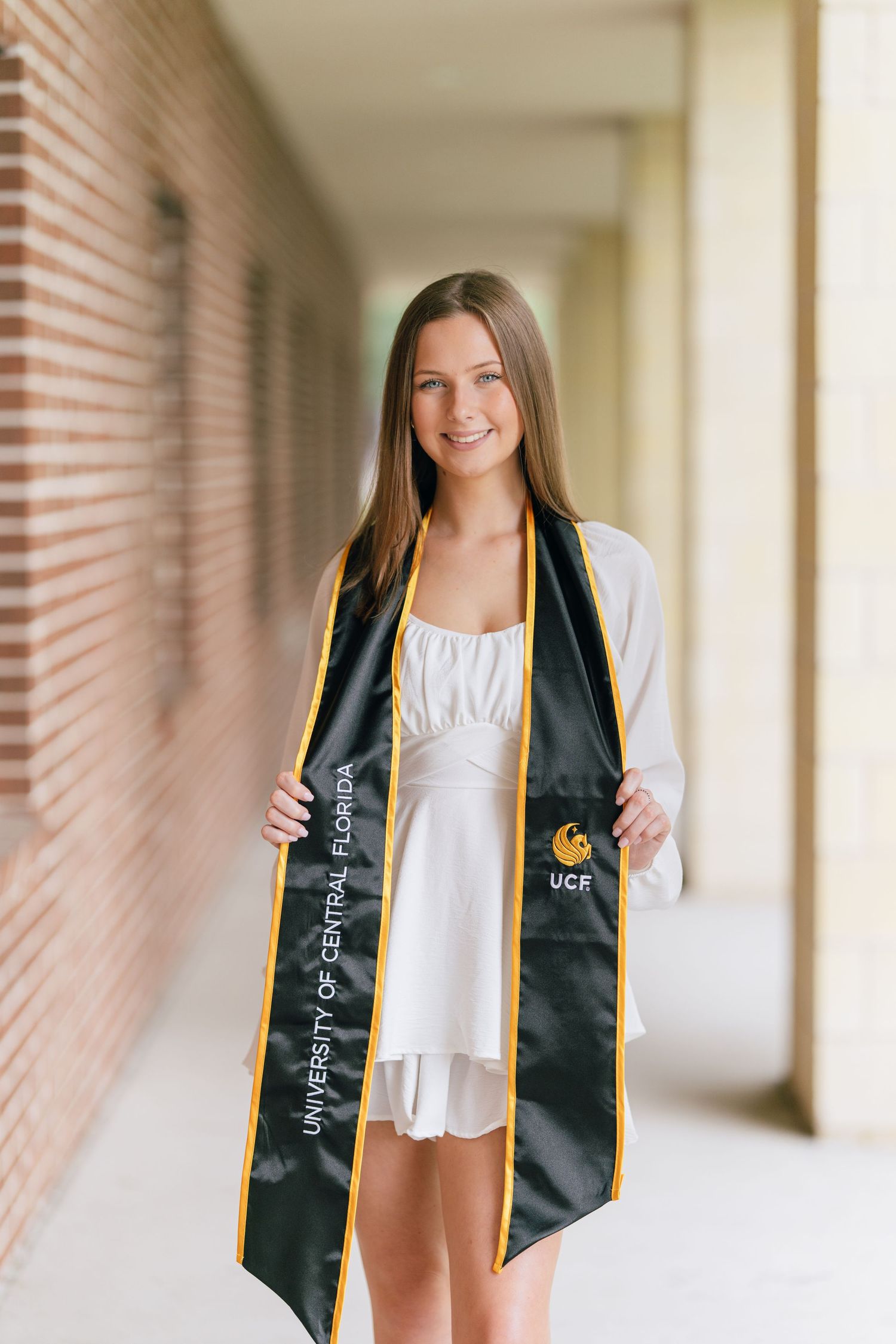 College graduate wearing white dress and black graduation stole with gold trim stands smiling in hallway corridor.