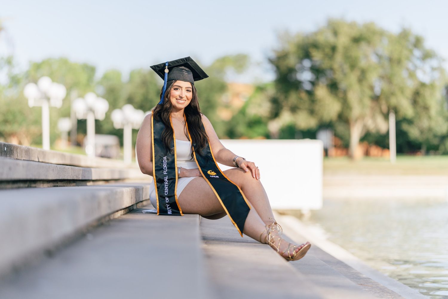 A smiling graduate in cap and gown sits on steps by a waterfront, celebrating her academic achievement outdoors.